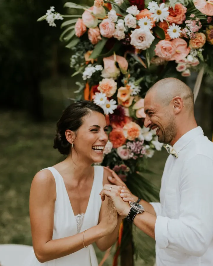 Jeunes mariés éclatant de rire sous une arche fleurie le jour de leur mariage