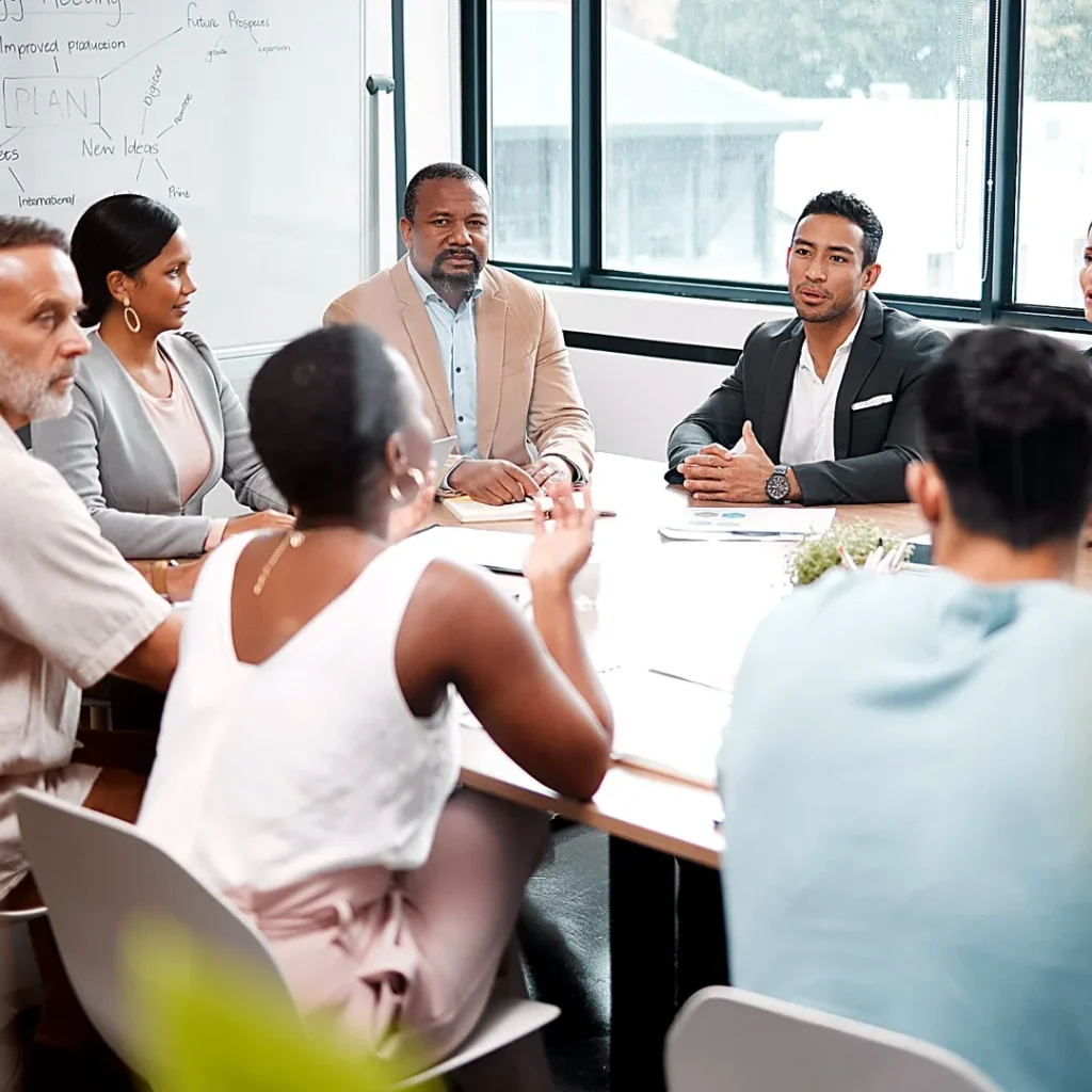 Des professionnels réunis autour d’une grande table de réunion échangent devant un tableau blanc.