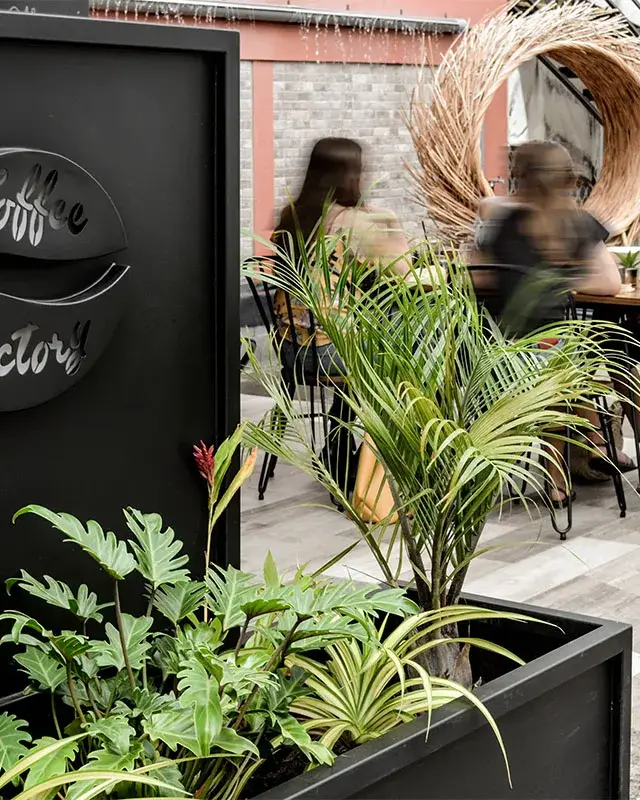 Terrasse du coffee bar avec jardinières noires plantées, logo en métal découpé et cercle en fibres naturelles