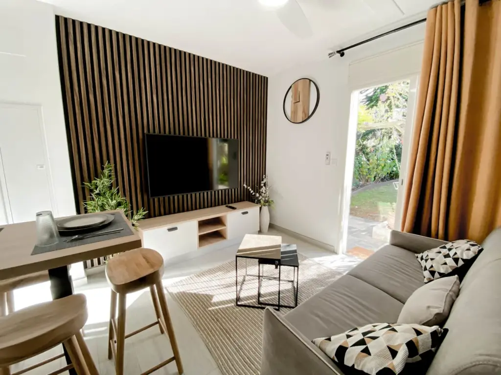 Séjour d’un appartement en location saisonnière au Tampon : mur en tasseaux de bois foncé, canapé gris, coin repas avec tabourets en bois et porte-fenêtre sur le jardin