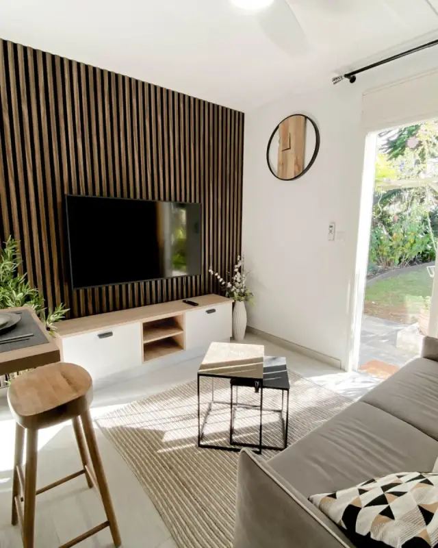 Séjour d’un appartement en location saisonnière au Tampon : mur en tasseaux de bois foncé, canapé gris, coin repas avec tabourets en bois et porte-fenêtre sur le jardin