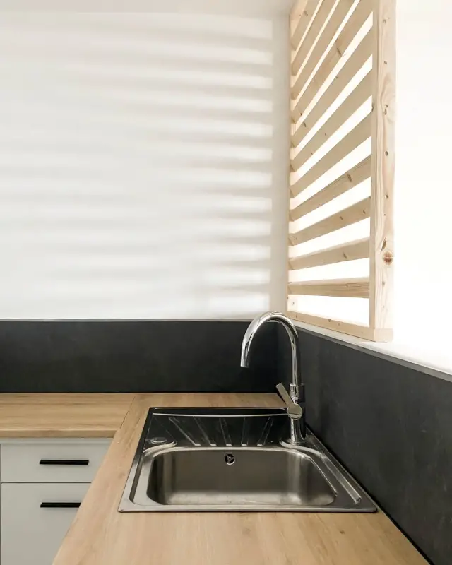 Cuisine blanche avec crédence noire, plan effet chêne et claustra à lames horizontales en pin devant la fenêtre