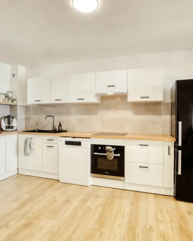 Cuisine en L blanche aux poignées noires, plan effet chêne, crédence beige et réfrigérateur noir