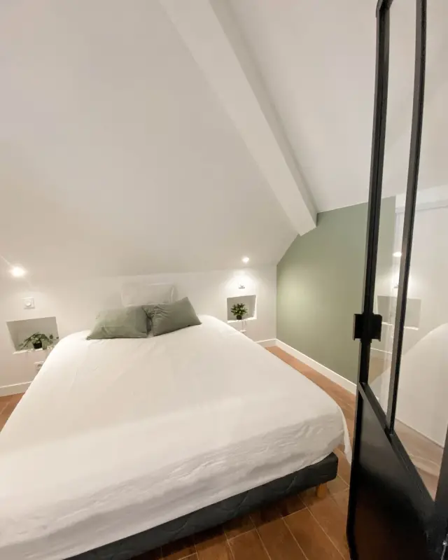 Chambre sous les combles avec mur vert sauge, niches éclairées en chevets et porte vitrée noire