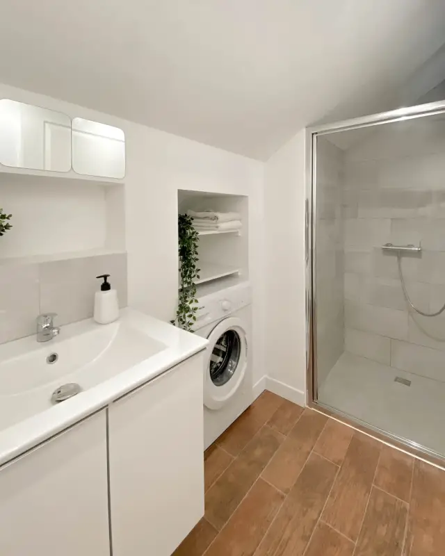 Salle d’eau blanche avec meuble vasque, niche pour le lave-linge, étagères à linge et douche vitrée