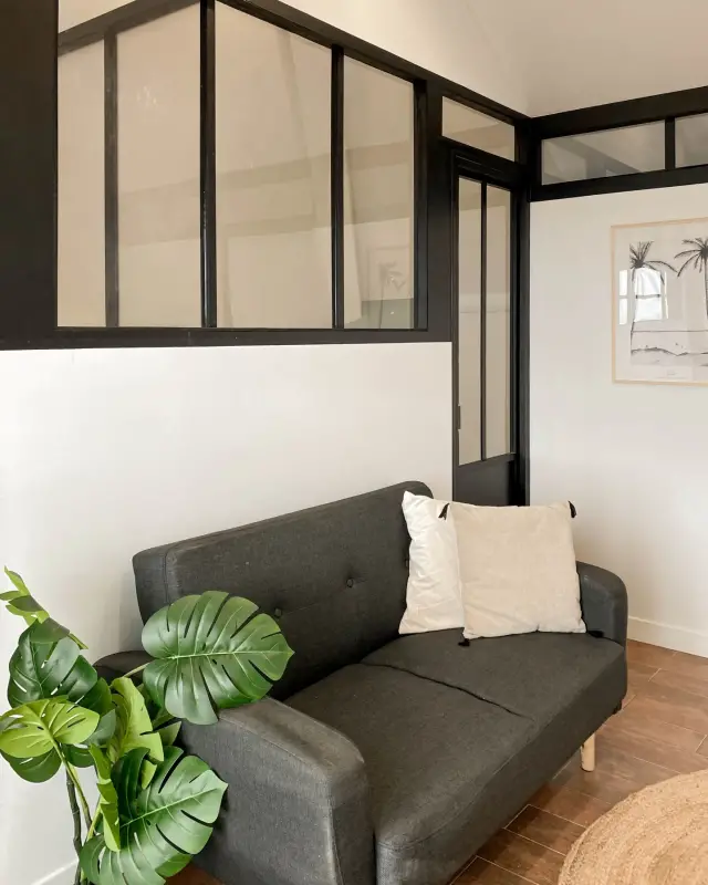 Verrière d’atelier noire au-dessus d’un canapé anthracite, monstera et tapis de jute rond