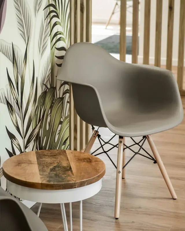 Fauteuil coque gris et guéridon en bois devant un papier peint à feuillages et un claustra