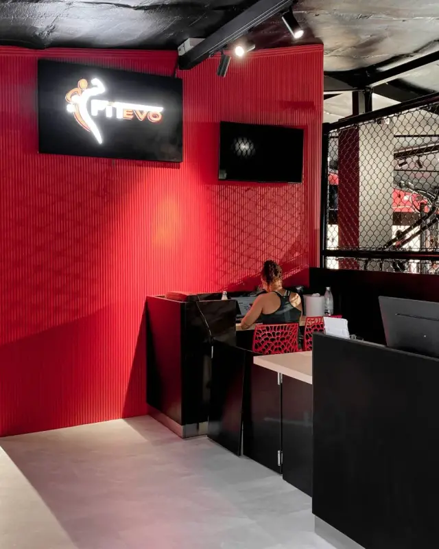 Accueil de la salle de sport avec volume rouge rainuré, enseigne lumineuse, banque noire et cloison en grillage