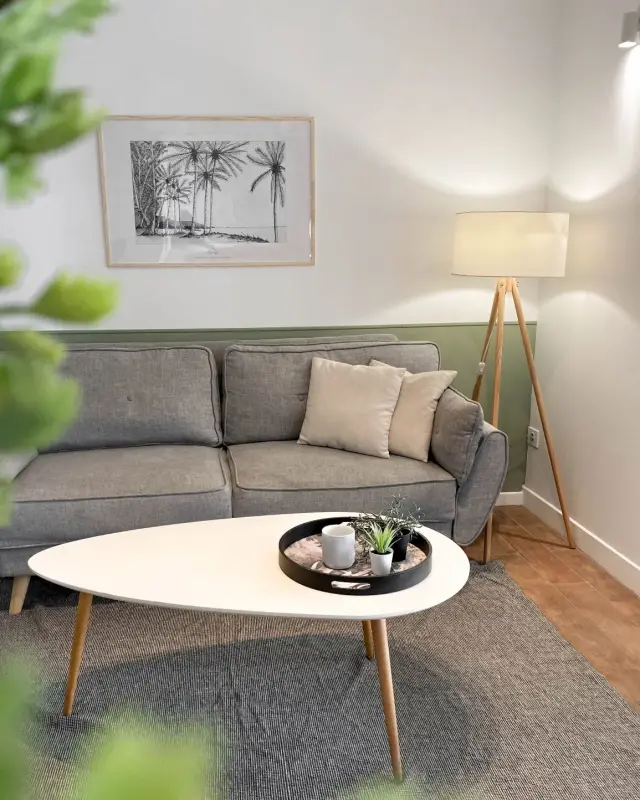 Séjour d’un des appartements créés à L’Étang-Salé avec canapé gris, soubassement vert sauge, affiche de palmiers et lampadaire trépied