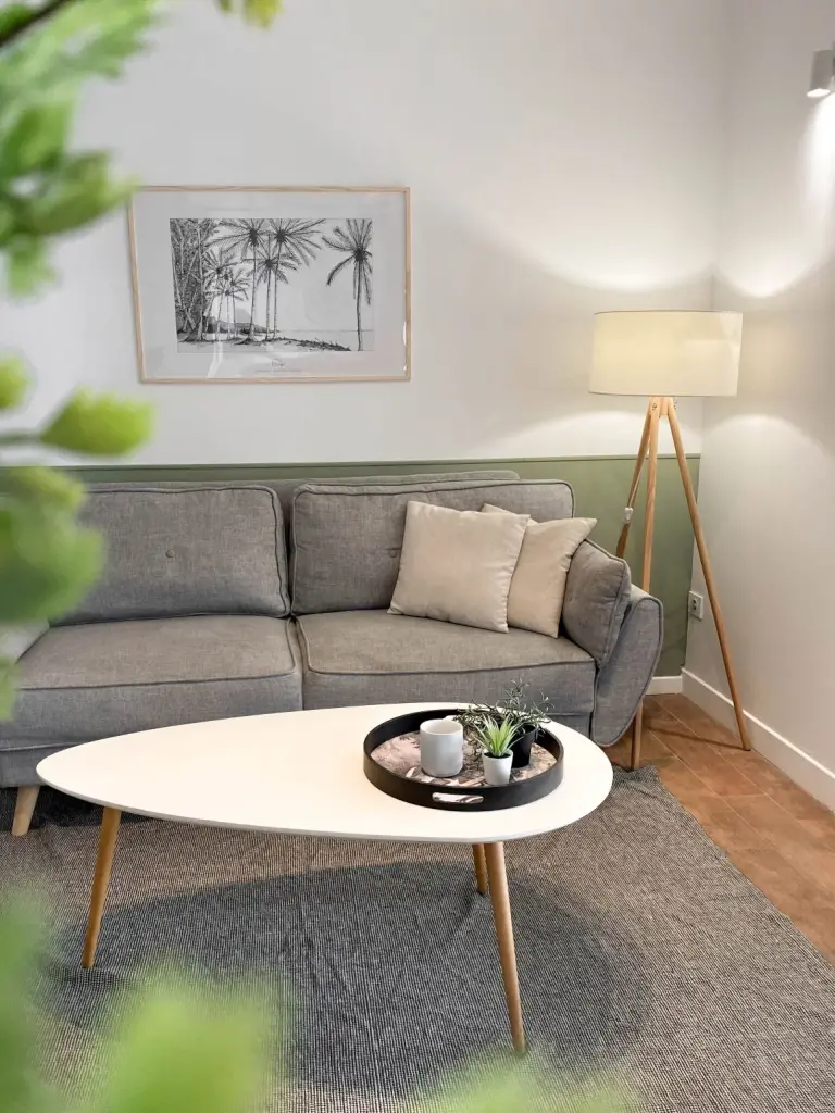 Séjour d’un des appartements créés à L’Étang-Salé avec canapé gris, soubassement vert sauge, affiche de palmiers et lampadaire trépied