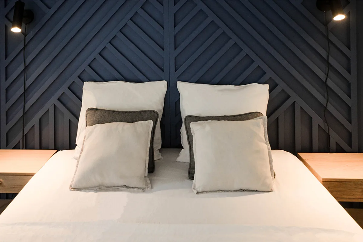 Chambre d’une location saisonnière à Saint-Gilles-les-Bains : tête de lit en tasseaux géométriques bleu nuit, appliques noires et chevets en bois
