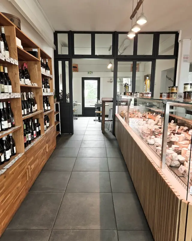 Allée entre rayonnages de vins en chêne massif et vitrine de fromages, verrière noire au fond