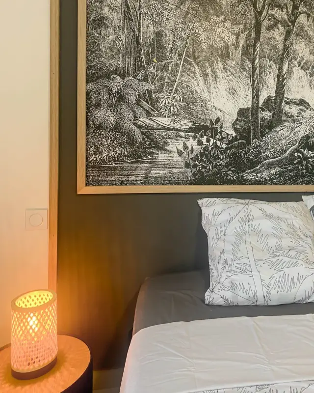 Chambre au mur gris foncé avec grand cadre de palmiers gravés, lampe en bambou tressé et linge imprimé palmiers