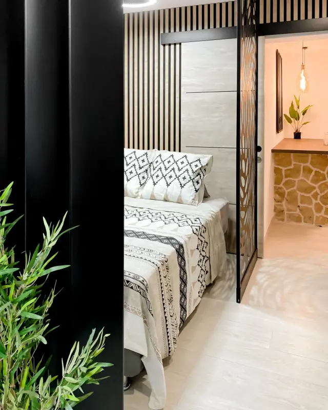 Chambre avec tête de lit en tasseaux de bois clair, parure noire et blanche et paravent en métal ajouré