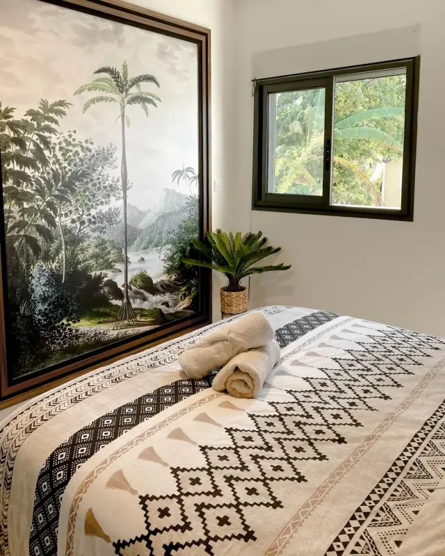 Chambre avec grand tableau de paysage tropical, cycas en pot et serviettes pliées sur le lit
