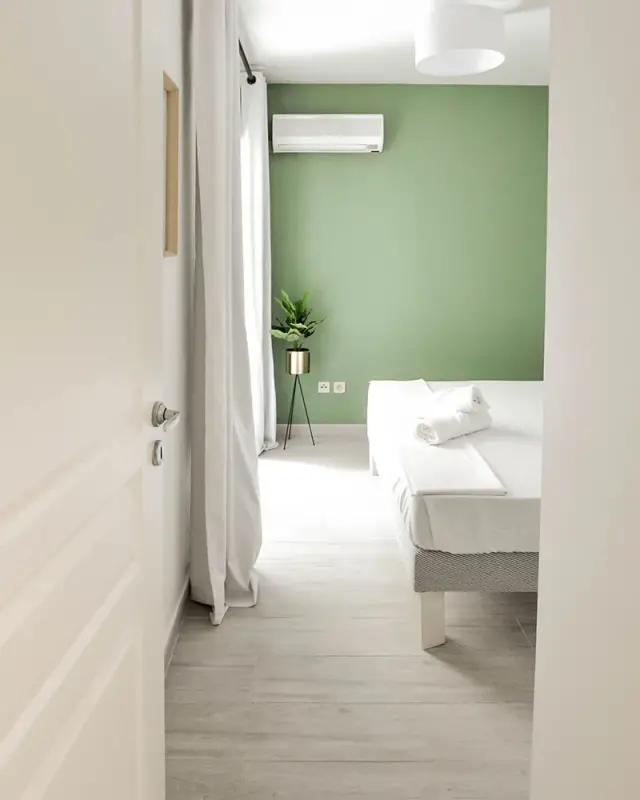 Chambre avec mur vert sauge, lit blanc, plante sur trépied doré et rideaux blancs
