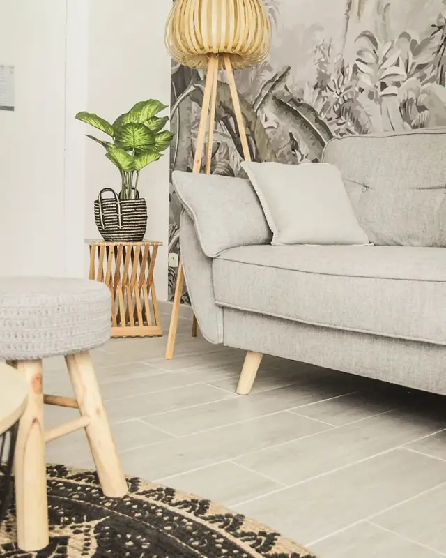 Pouf tricoté sur pieds en bois, tapis rond noir et naturel et canapé gris devant le papier peint tropical