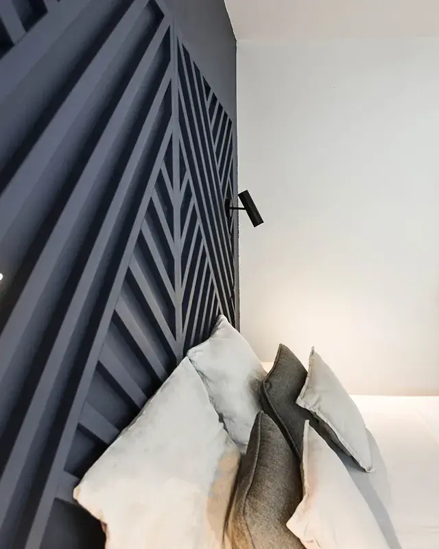Liseuse murale noire au premier plan, lit et coussins devant le mur bleu nuit