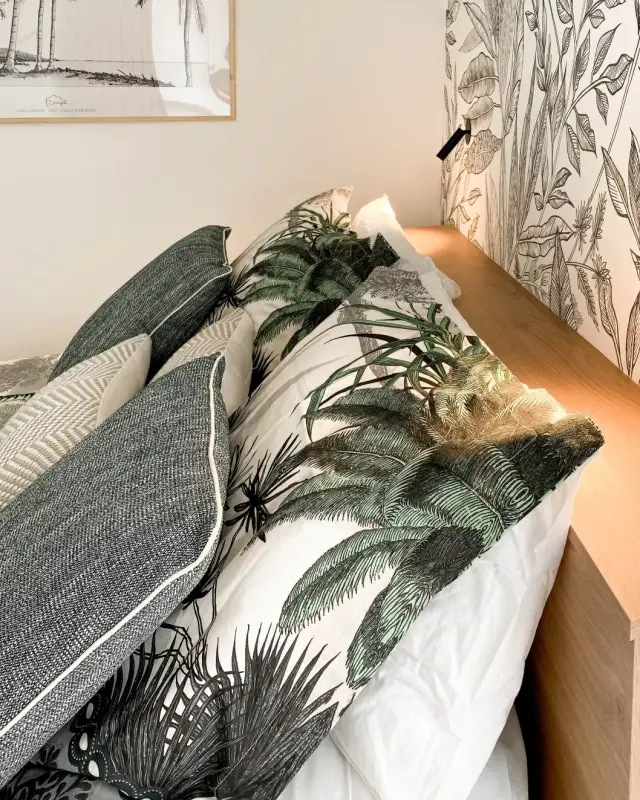 Tête de lit en bois éclairée, coussins à motifs de palmiers, papier peint à feuillages et affiche Lign lorizon