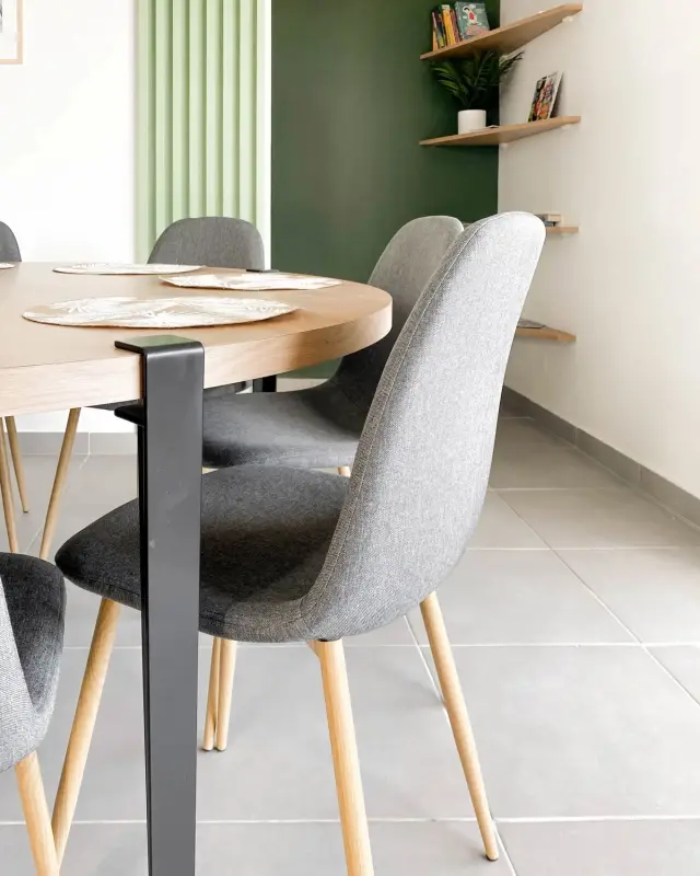 Chaises en tissu gris autour d’une table en bois sur pieds noirs, étagères d’angle en bois sur un mur vert