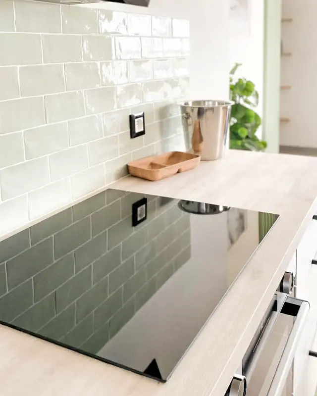 Plaque de cuisson noire devant une crédence en carreaux vert céladon, espace repas en arrière-plan