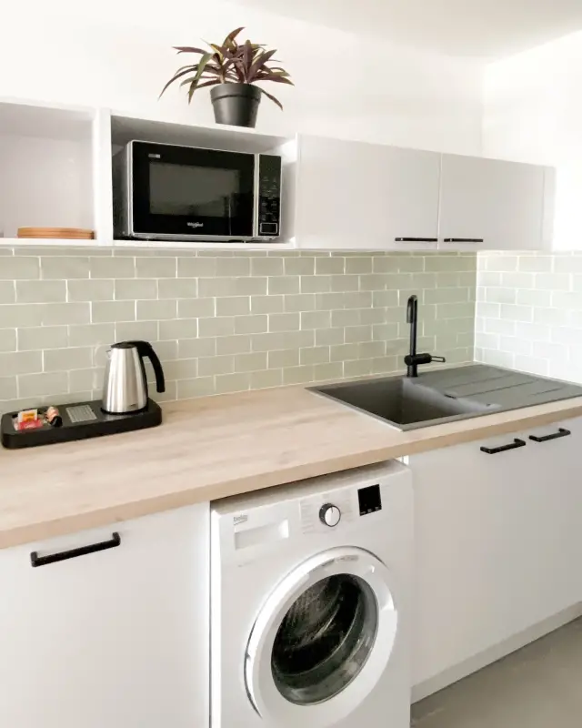 Cuisine blanche avec plan de travail effet bois, crédence en carreaux métro vert céladon, évier noir et lave-linge intégré