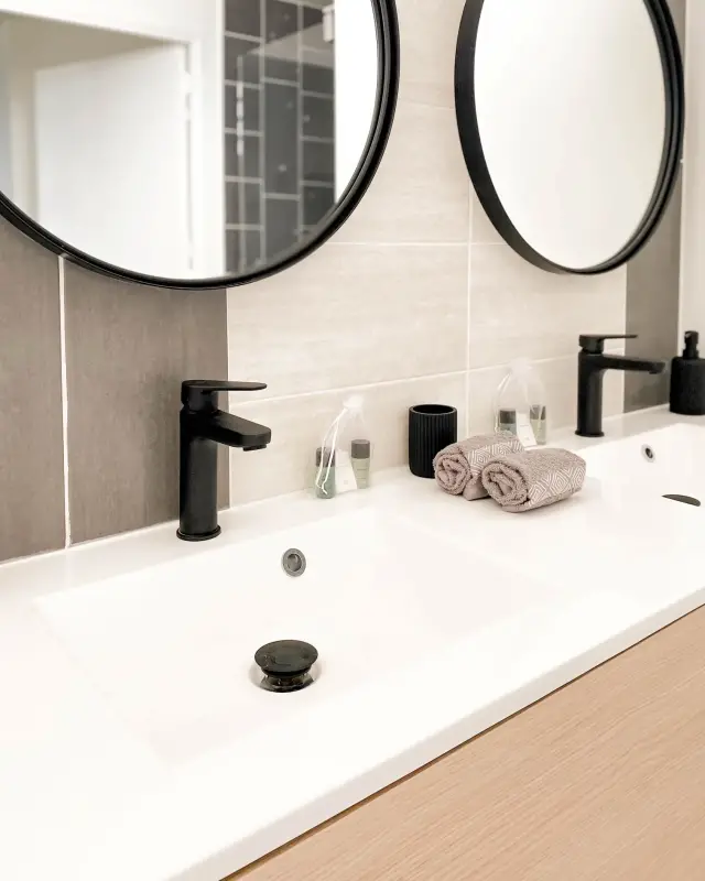 Double vasque blanche sur meuble en bois, miroirs ronds cerclés de noir et robinets noirs