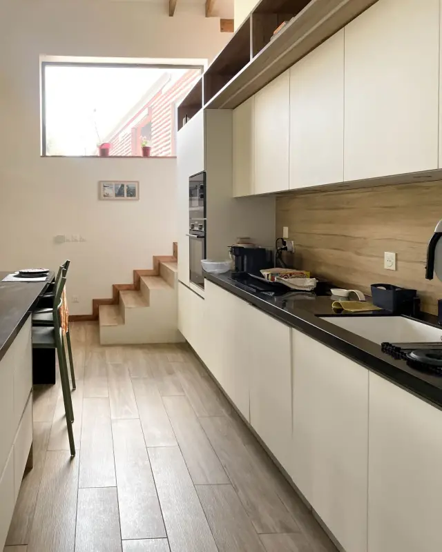 Cuisine linéaire aux façades crème, plan de travail noir, crédence effet bois et niches ouvertes