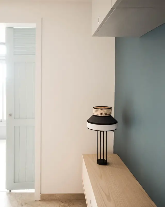 Meuble bas en bois clair et lampe en cannage devant un mur bleu profond, porte à persiennes ouverte