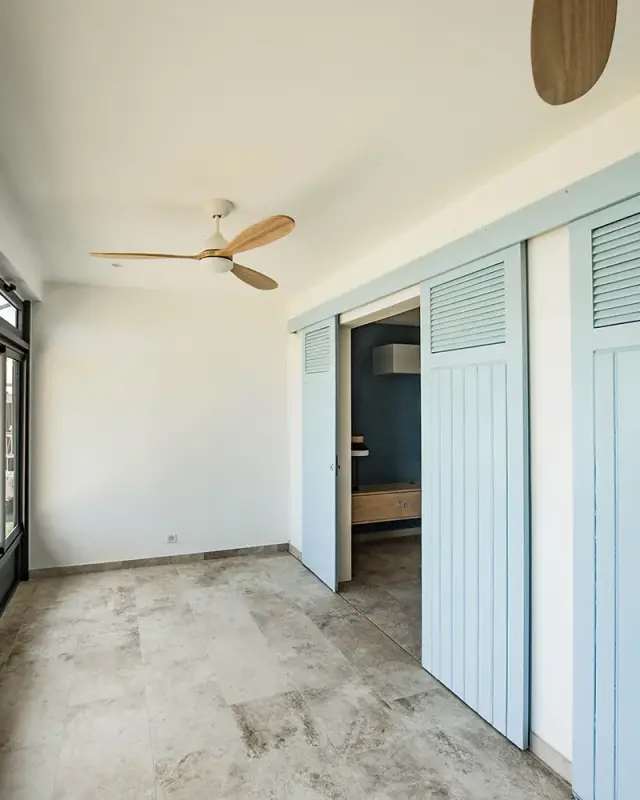 Pièce lumineuse avec portes coulissantes à persiennes bleu ciel, ventilateur à pales en bois et sol effet pierre