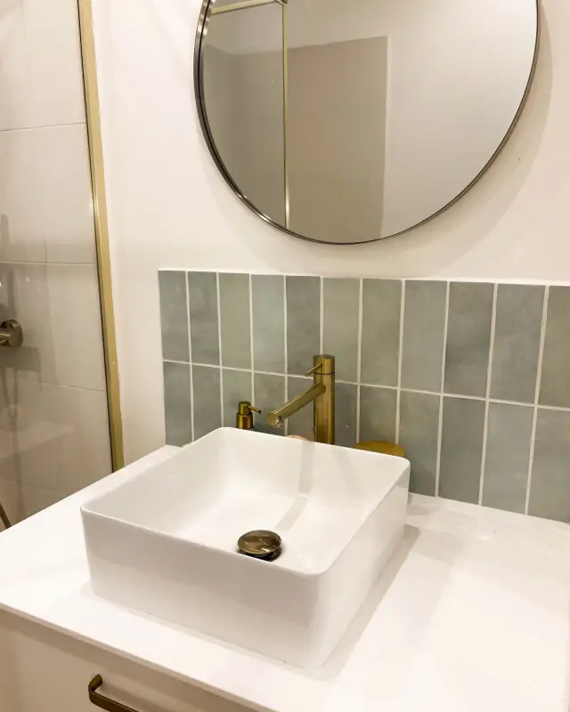 Salle d’eau avec vasque carrée à poser, robinetterie en laiton, miroir rond et crédence vert sauge