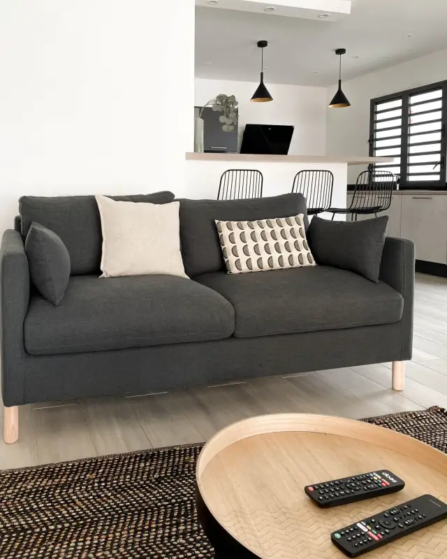 Canapé anthracite avec coussins, table basse ronde en bois et tapis tressé, cuisine en arrière-plan