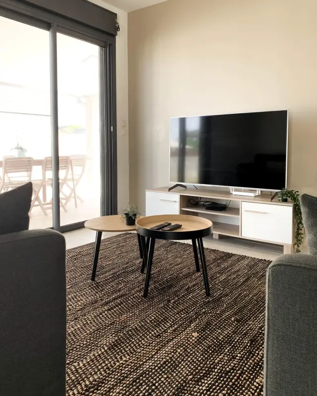 Salon avec tables gigognes noire et bois, tapis tressé, meuble TV et baie ouverte sur la terrasse